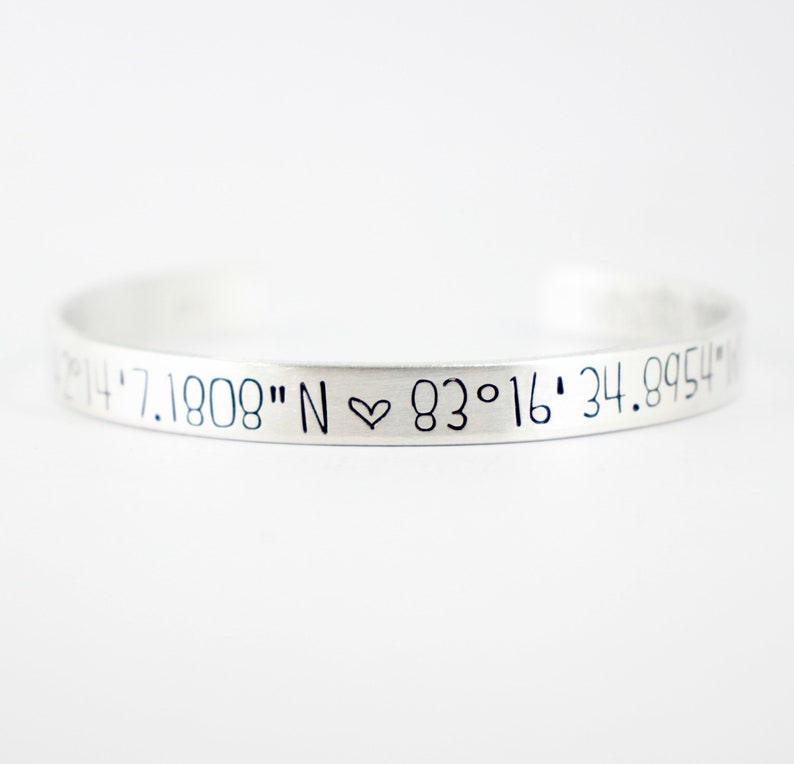May include: A silver-colored metal cuff bracelet featuring engraved black text that reads 42&deg;14'7.1808" N ♡ 83&deg;16'34.8954" N. The bracelet has a smooth, matte finish and a simple, open-ended circular design that curves gently across the frame.