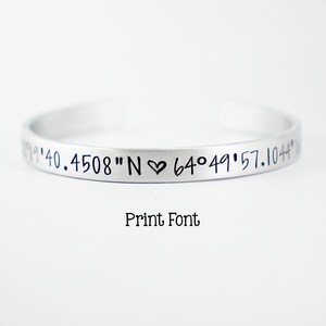 May include: A silver-toned metal cuff bracelet featuring engraved black text that reads 19'40.4508"N heart 64&deg;49'57.1044". Below the bracelet, the text Print Font is displayed in a simple black typeface against a plain white background.