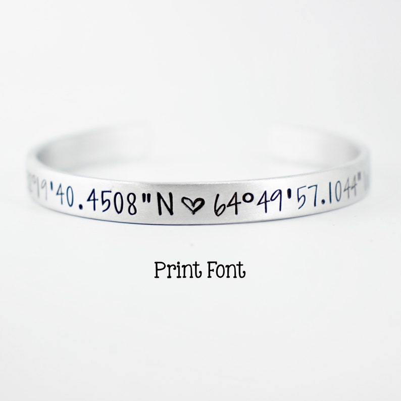 May include: A silver-toned metal cuff bracelet featuring engraved black text that reads 19'40.4508"N heart 64&deg;49'57.1044". Below the bracelet, the text Print Font is displayed in a simple black typeface against a plain white background.