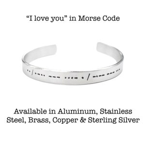May include: A silver cuff bracelet engraved with the Morse code for "I love you." The open-ended bracelet is made of metal and features the dot-dash pattern of Morse code. The text "I love you" in Morse Code is above the bracelet.