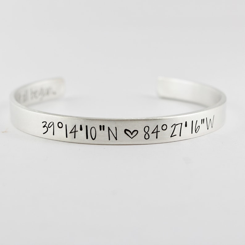 May include: A silver-toned metal cuff bracelet featuring engraved black text that reads 39&deg;14'10"N ♡ 84&deg;27'16"W on the outer surface. The bracelet has an open-ended design and includes additional engraved text on the inner surface that reads it all began.