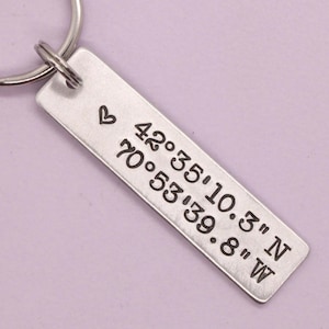 May include: A silver-toned rectangular metal keychain tag featuring a stamped heart symbol and the engraved coordinates 42°35'10.3" N 70°53'39.8" W. The tag is attached to a circular metal split ring, showcasing a clean, brushed metallic finish.