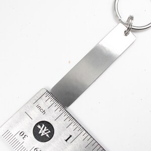 May include: A silver metal keychain with a blank rectangular tag measuring 1 inch in length. The tag is attached to a silver metal key ring.