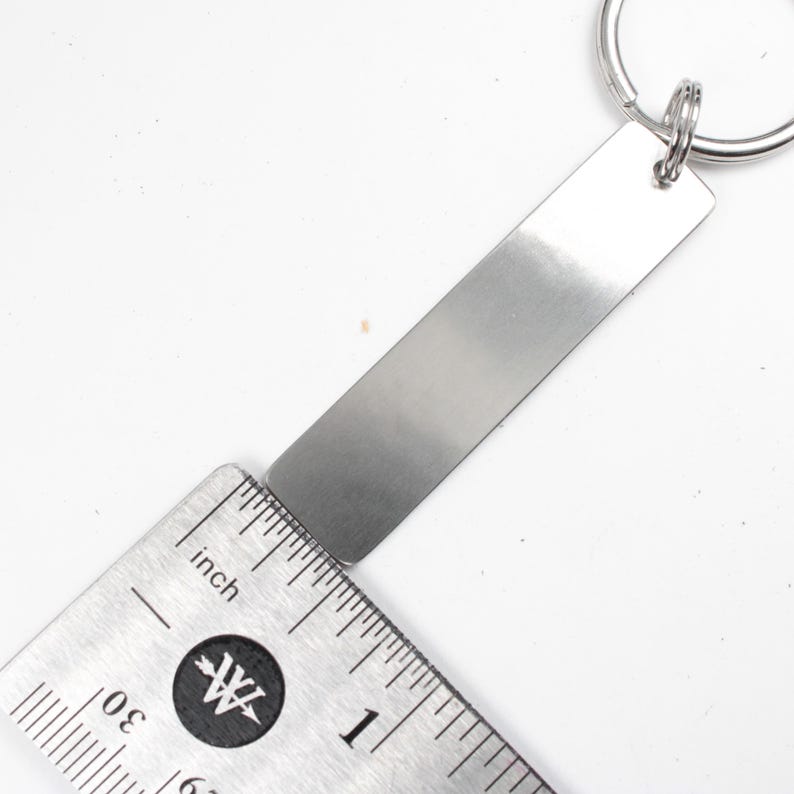 May include: A silver metal keychain with a blank rectangular tag measuring 1 inch in length. The tag is attached to a silver metal key ring.
