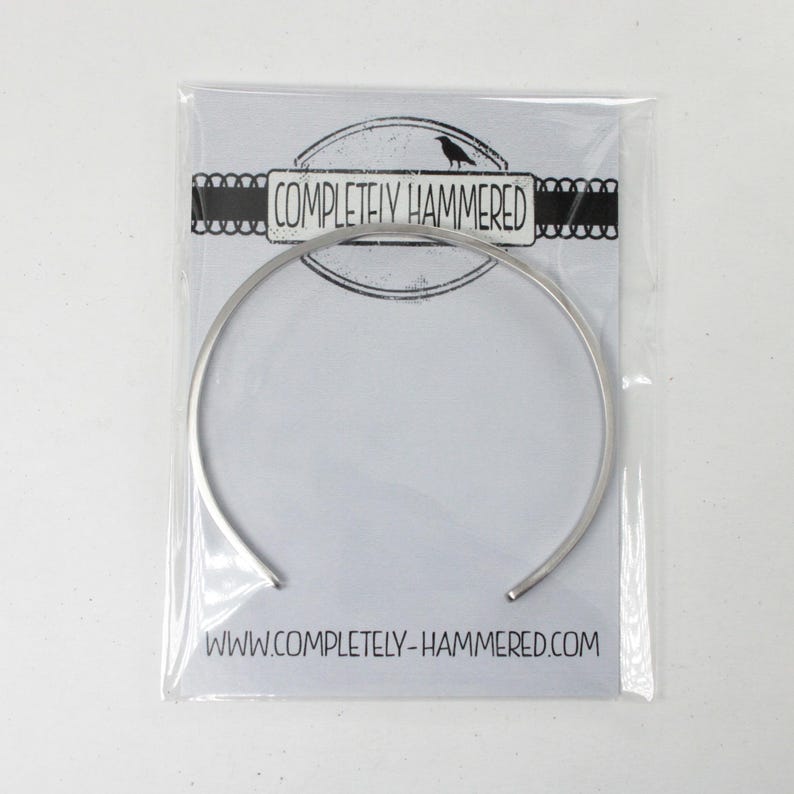 May include: A silver-toned metal cuff bracelet with a smooth, thin band. The bracelet is packaged in a clear plastic sleeve on a white card featuring a black raven logo, the text "COMPLETELY HAMMERED," and the website URL "WWW.COMPLETELY-HAMMERED.COM."