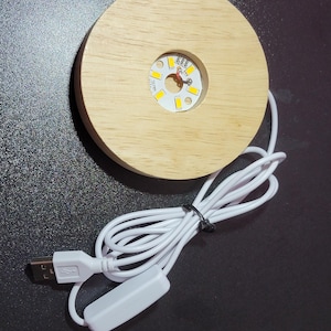 May include: A round, light-colored wooden LED light base with a circular cutout for the light. The base has a white USB cable with an on/off switch. The light is a warm white color.