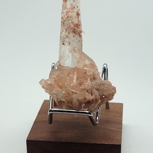 May include: A clear quartz crystal with red inclusions, mounted on a wooden base with a metal stand.