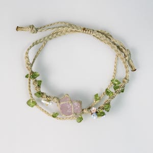 以下が含まれることがあります： ピンクのクリスタルチャーム、緑の葉、小さな白と青の花が付いた手作りブレスレット。ブレスレットは天然の紐で作られています。