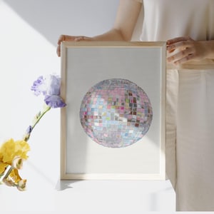 May include: A framed print featuring a colorful disco ball design. The artwork showcases a mosaic of small, reflective squares in shades of pink, blue, and silver. The print is held by a person, with a flower in the foreground.