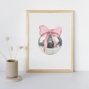 Puede incluir: Impresión enmarcada de una bola de discoteca plateada con un lazo rosa. La bola de discoteca tiene una superficie reflectante, y el lazo es de color rosa suave. El marco es de madera clara.