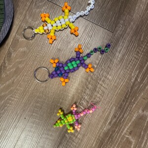 May include: Three beaded lizard keychains. One is white with orange accents, one is purple with green accents, and one is green with pink accents.