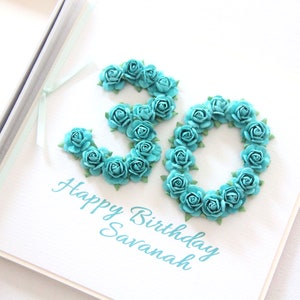 May include: A white birthday card with the number 30 formed from teal paper roses. The words "Happy Birthday Savanah" are written in a matching teal script below the number. The card is open, revealing a light blue ribbon.