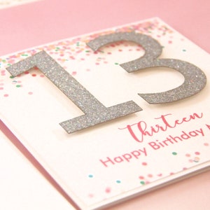 May include: A white card with silver glitter numbers "13" and the text "Thirteen Happy Birthday". The background is pink with confetti.