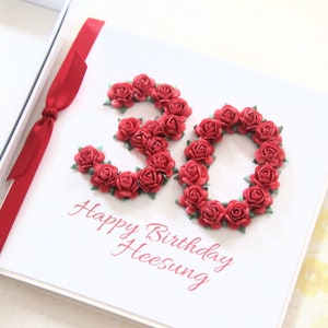 May include: A white birthday card with the number 30 formed from red paper roses. The words "Happy Birthday Heesung" are printed in red script below the number. A red ribbon is tied to the left side of the card.