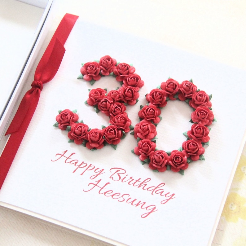 May include: A white birthday card with the number 30 formed from red paper roses. The words "Happy Birthday Heesung" are printed in red script below the number. A red ribbon is tied to the left side of the card.