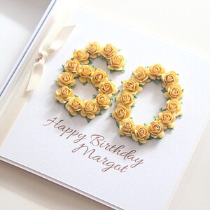 May include: A white birthday card with the number 60 formed by yellow roses. The words "Happy Birthday Margot" are written in brown script below the floral arrangement. A cream ribbon is tied to the left side.