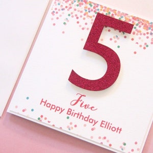 May include: A white card with a pink glitter number five and confetti. The card says "Five Happy Birthday Elliott".