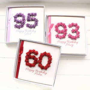 May include: Three birthday cards in white boxes. Each card features a number made of small roses. The numbers are 95 in purple, 93 in pink, and 60 in red. Each card has a ribbon and a birthday message.