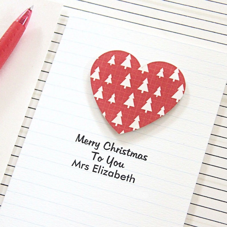 May include: A red heart-shaped card with a white Christmas tree pattern. The card has the text "Merry Christmas To You Mrs Elizabeth" written on it.