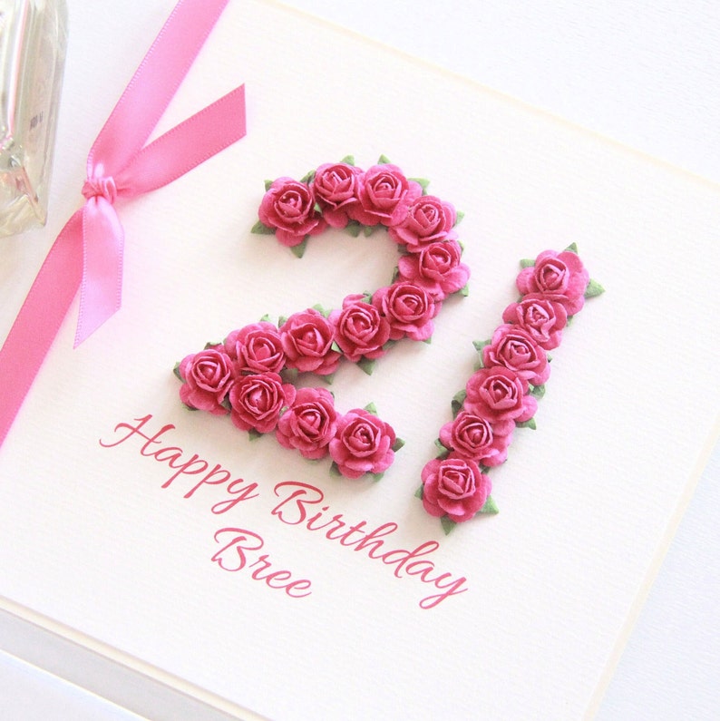 May include: A handmade birthday card with the number 21 formed from pink roses. The words "Happy Birthday Bree" are written in pink script below the floral arrangement. A pink ribbon is tied to the left side of the card.
