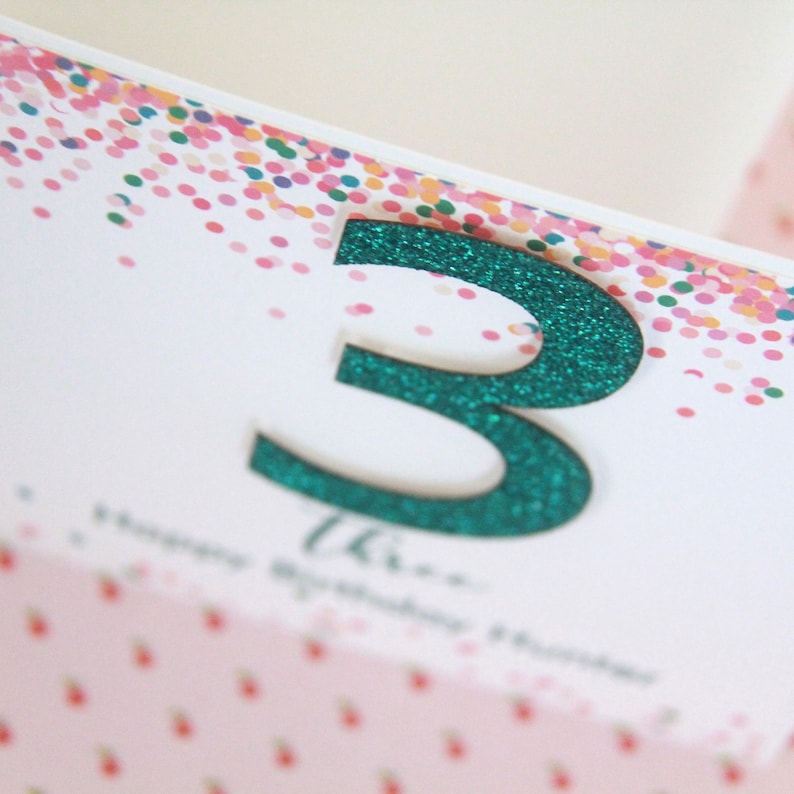 May include: A white card with a green glitter number three on it. The card has a confetti pattern in the background.