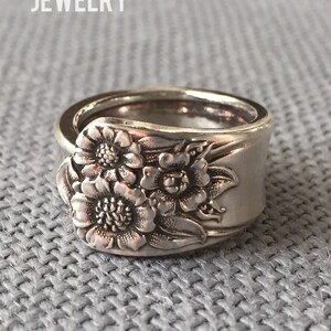 May include: A silver spoon ring with a floral design. The ring is made from a vintage spoon and features three flowers with detailed carvings. The ring is a unique and handmade piece of jewelry.
