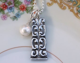 May include: A silver necklace with a silver pendant featuring a decorative design and a white pearl charm. The necklace is on a white background with a floral pattern.