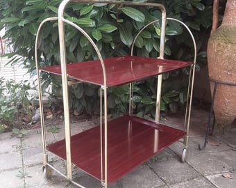 May include: A two-tiered serving cart with a gold metal frame and burgundy wood shelves. The cart has four wheels and a handle on the top.