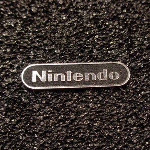 Könnte beinhalten: Ein silbernes Metalletikett mit dem Wort "Nintendo" in schwarzen Buchstaben.