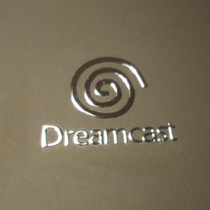 Könnte beinhalten: Ein silbernes Dreamcast-Logo mit einem stilisierten Wirbeldesign auf beigefarbenem Hintergrund.