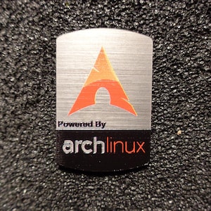 Peut inclure: Un autocollant en métal argenté avec un logo Arch Linux rouge et orange et le texte "Powered By archlinux".