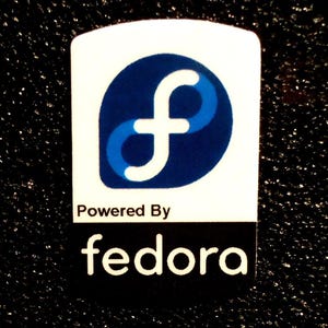 Puede incluir: Una pegatina rectangular con fondo blanco, que presenta un símbolo 'f' estilizado en azul y blanco dentro de un círculo azul. Debajo del símbolo, el texto dice 'Powered By' sobre la palabra 'fedora' en blanco sobre un rectángulo negro.
