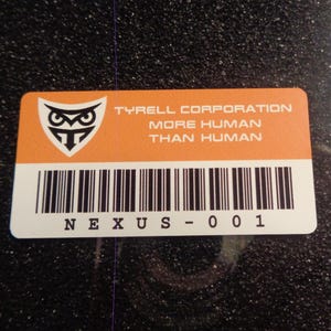 May include: An orange and white rectangular sticker with the text "TYRELL CORPORATION MORE HUMAN THAN HUMAN" and a barcode. The sticker also features a logo of an owl and the text "NEXUS - 001".