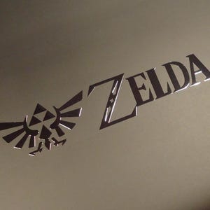 Può includere: Un logo metallico argentato con la parola "ZELDA" in un font stilizzato. Il logo è su uno sfondo marrone chiaro e presenta un uccello stilizzato con le ali spiegate.