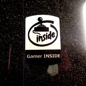 Puede incluir: Una pegatina blanca con un icono de joystick negro y la palabra "inside" en un círculo. Debajo del círculo está el texto "Gamer INSIDE".