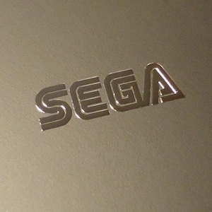 Peut inclure: Un logo métallique argenté avec le mot "SEGA" dans une police de caractères stylisée.