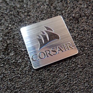 Puede incluir: Una insignia cuadrada de metal plateado con un logotipo de Corsair negro y la palabra "CORSAIR" en letras negras.