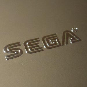 Op de afbeelding: Een zilveren, gegraveerd SEGA-logo op een lichtbruin oppervlak.