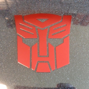May include: A red Transformers logo with a stylised face, featuring a triangular forehead, two horizontal lines for eyebrows, and a rectangular mouth.