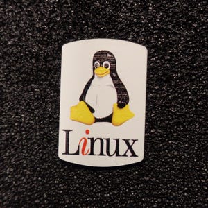 Peut inclure: Un autocollant blanc avec une illustration de pingouin noir et blanc et le mot "Linux" en lettres rouges et noires.