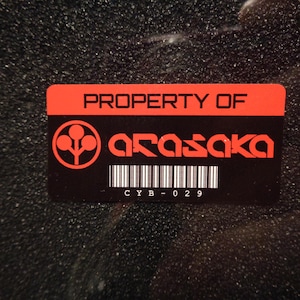 Puede incluir: Pegatina negra y roja con el texto "PROPERTY OF Arasaka" y un código de barras con el texto "CYB-029".
