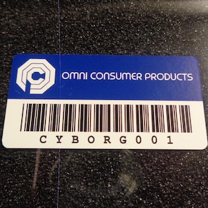 May include: A white and blue rectangular label with a black and white barcode. The label has a black and white logo with the letter "C" inside a circle. The text "OMNI CONSUMER PRODUCTS" is printed in white on a blue background. The text "CYBORG 001" is printed in black below the barcode.
