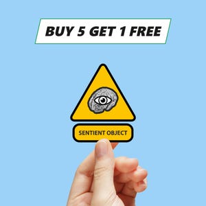 Puede incluir: Un cartel de advertencia amarillo con borde negro y un cerebro gris con un ojo en su interior. El cartel dice "Objeto Sensible" y tiene el texto "COMPRA 5, LLEVA 1 GRATIS" encima.