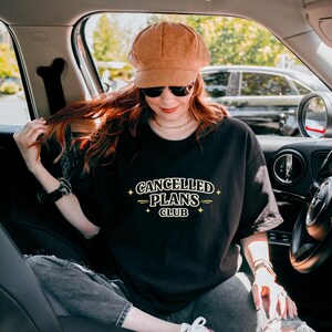 Puede incluir: Camiseta negra con el texto "CANCELLED PLANS CLUB" en fuente blanca estilizada. La camiseta se usa con una gorra de pana marr&oacute;n y gafas de sol. La persona est&aacute; sentada en un coche.
