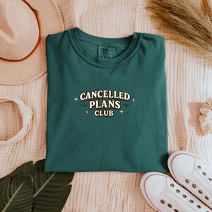 Puede incluir: Una camiseta verde oscuro con el texto "CANCELLED PLANS CLUB" en blanco, con una fuente retro. La camiseta est&aacute; doblada y descansa sobre una superficie color crema, con zapatillas blancas y un sombrero en el fondo.