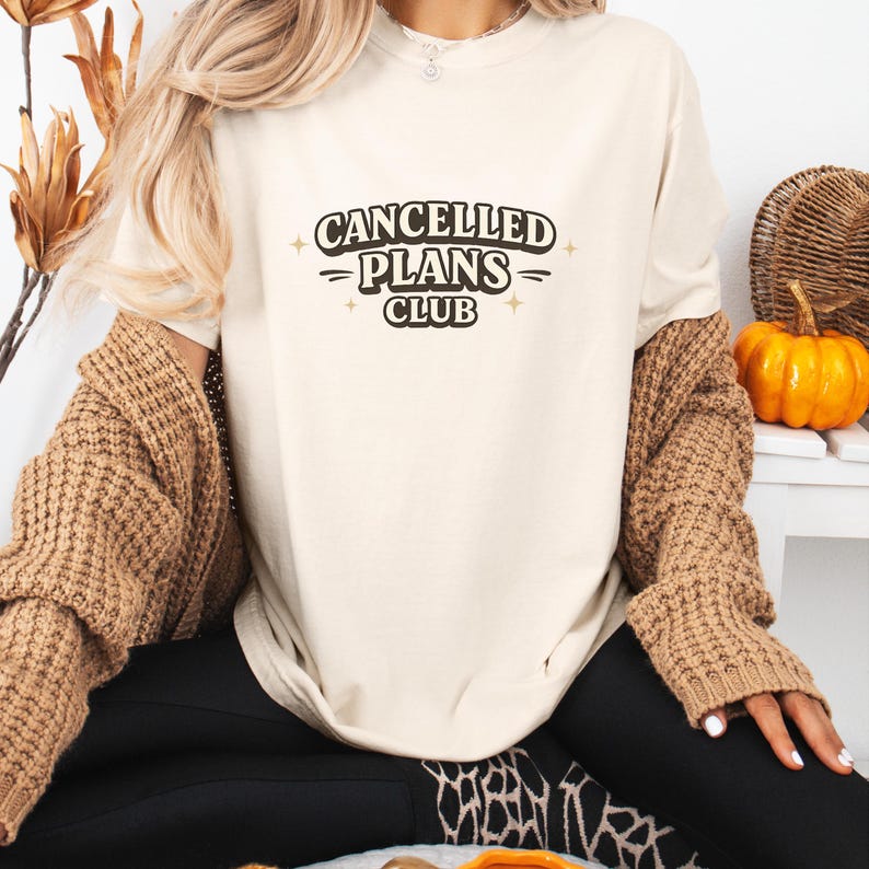 Puede incluir: Camiseta color crema con el texto "CANCELLED PLANS CLUB" en negro. La camiseta tiene un corte holgado. La persona de la imagen lleva una rebeca marr&oacute;n y pantalones negros. Una peque&ntilde;a calabaza y objetos decorativos est&aacute;n en el fondo.
