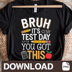 Op de afbeelding: Zwart T-shirt met witte tekst die "Bruh it's test day you got this" zegt. De tekst is omringd door gele en rode potloodtekeningen. Rechts van de tekst staat een rode appel.