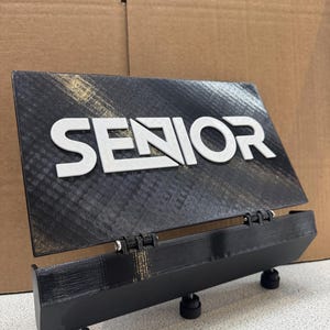 Può includere: Un cartello nero stampato in 3D con la parola "SENIOR" in lettere bianche. Il cartello è su un supporto nero con tre gambe regolabili.