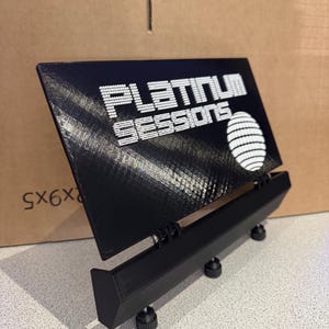 Peut inclure: Un panneau d'affichage rectangulaire noir avec le texte blanc "PLATINUM SESSIONS" et un globe graphique blanc. Le panneau est monté sur un support noir réglable avec trois boutons circulaires à la base et un mécanisme à charnière pour incliner.