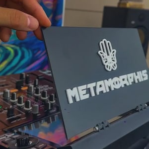 Pode incluir: Uma placa retangular cinza-escura com um ícone de mão hamsa branca em relevo acima do texto branco METAMORPHIS. A placa está posicionada em um ângulo, exibindo uma superfície plana e limpa com letras sem serifa em negrito e um símbolo.